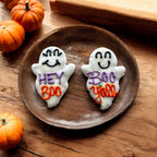 Two white ghost-shaped dog cookies with smiling faces and purple and orange lettering reading Hey Boo and Boo Y’all on a wooden plate.