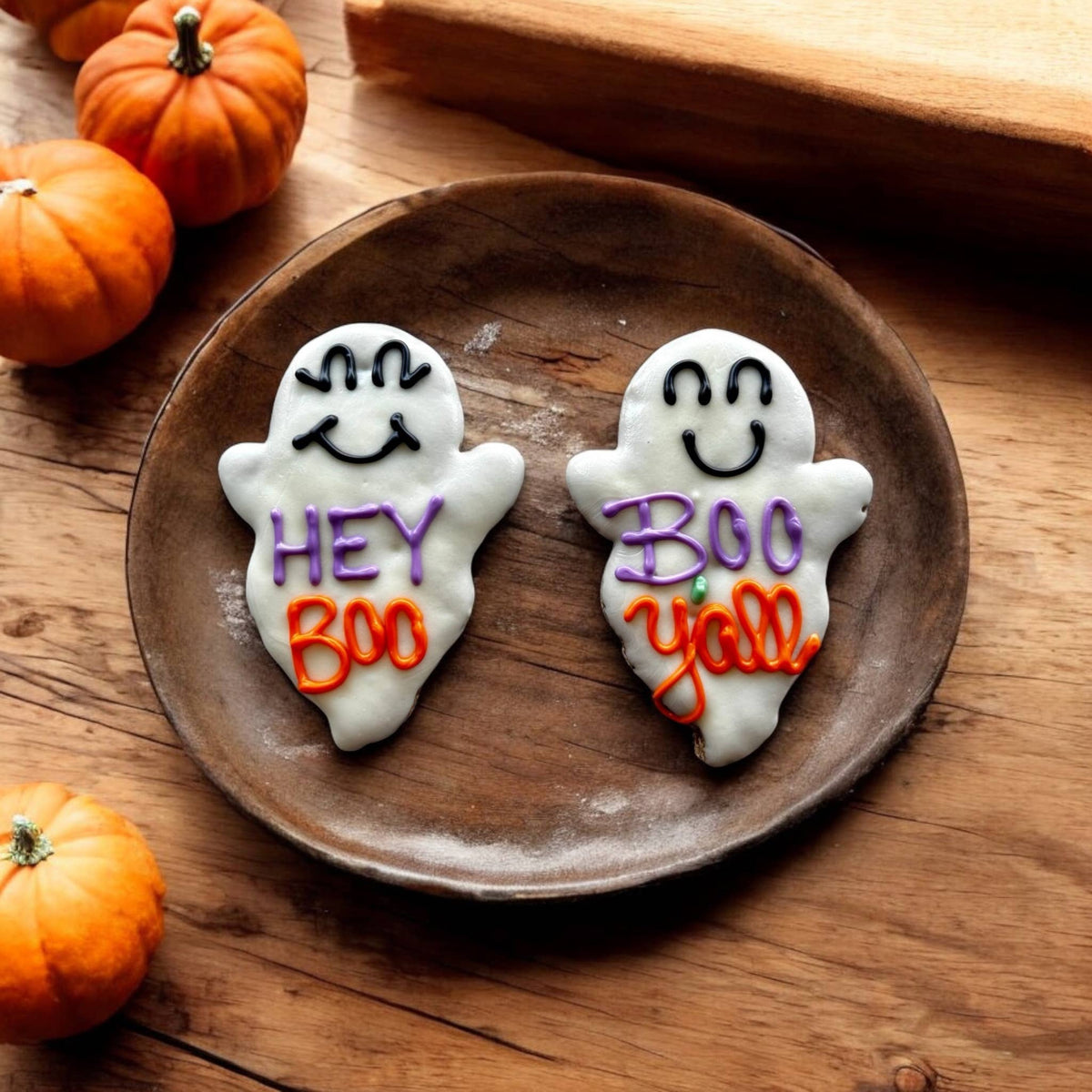 Two white ghost-shaped dog cookies with smiling faces and purple and orange lettering reading Hey Boo and Boo Y’all on a wooden plate.