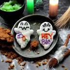 Two white ghost-shaped dog cookies with smiling faces and purple and orange lettering reading Hey Boo and Boo Y’all on a wooden plate.
