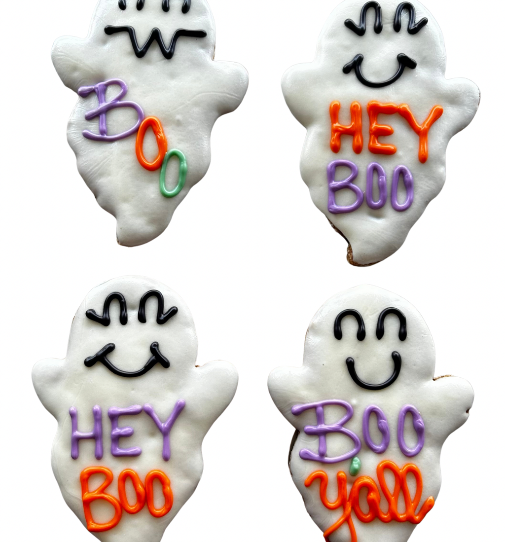 Two white ghost-shaped dog cookies with smiling faces and purple and orange lettering reading Hey Boo and Boo Y’all on a wooden plate.