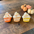 Three cupcake-shaped dog cookies with orange, tan, and yellow bases, white WOOF lettering, and white tops with fall sprinkles.