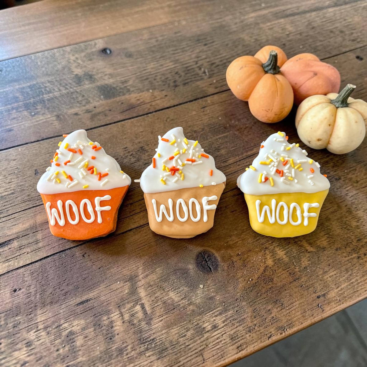 Three cupcake-shaped dog cookies with orange, tan, and yellow bases, white WOOF lettering, and white tops with fall sprinkles.