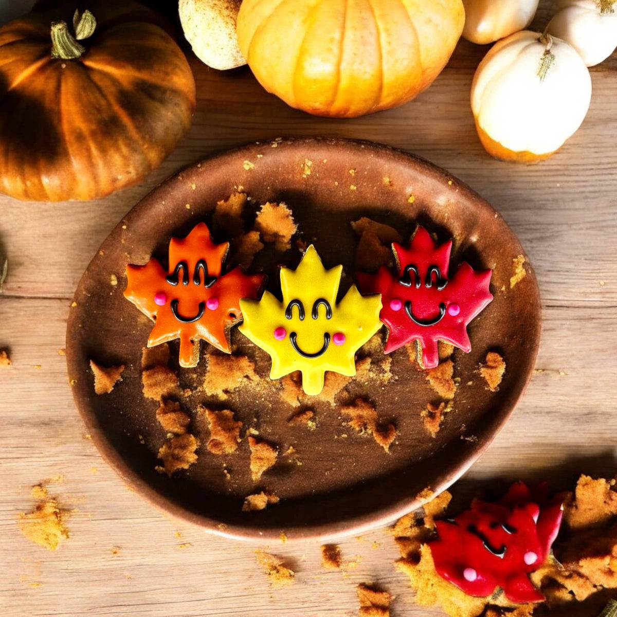 Orange, yellow, and red leaf-shaped dog cookies with smiling faces and pink cheeks on a rustic plate surrounded by pumpkins.