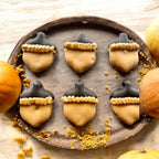 Six acorn-shaped dog cookies with tan decoration, dark caps, and piped trim arranged on a round gray plate.