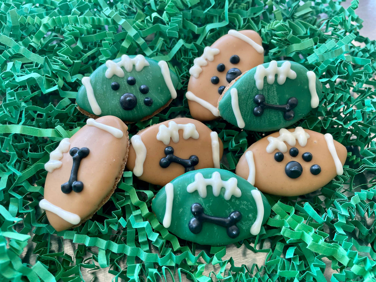 Green and tan football-shaped dog cookies decorated with white laces and black paw prints or bones on green shredded paper.