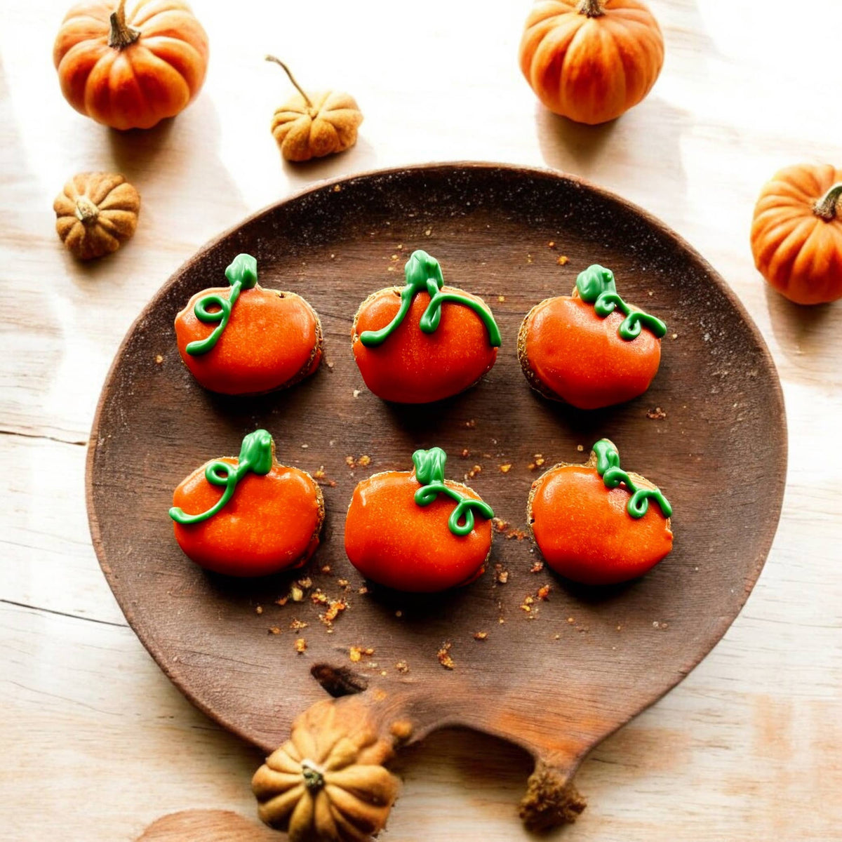 Six pumpkin-shaped dog cookies decorated in orange with green stems and curling vines on a round wooden tray.