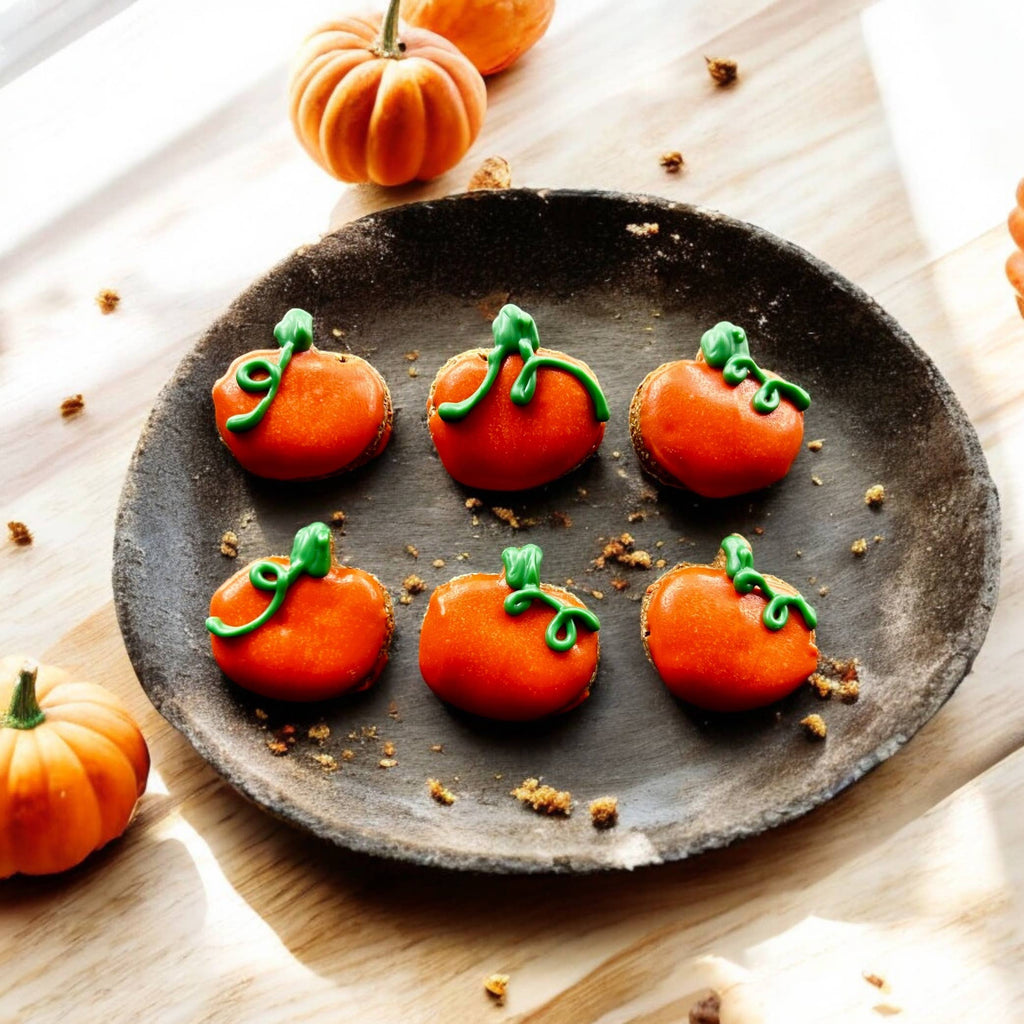 Six pumpkin-shaped dog cookies decorated in orange with green stems and curling vines on a round wooden tray.