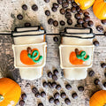 Two mini coffee-cup-shaped dog cookies with white decoration, black lid outlines, and orange pumpkins with green vines on a wooden surface.
