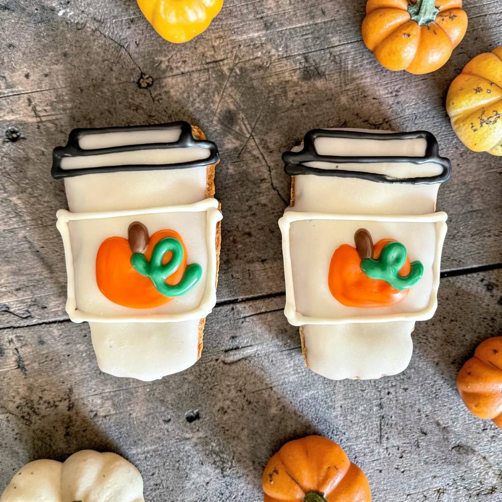 Two mini coffee-cup-shaped dog cookies with white decoration, black lid outlines, and orange pumpkins with green vines on a wooden surface.