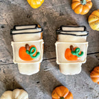 Two mini coffee-cup-shaped dog cookies with white decoration, black lid outlines, and orange pumpkins with green vines on a wooden surface.