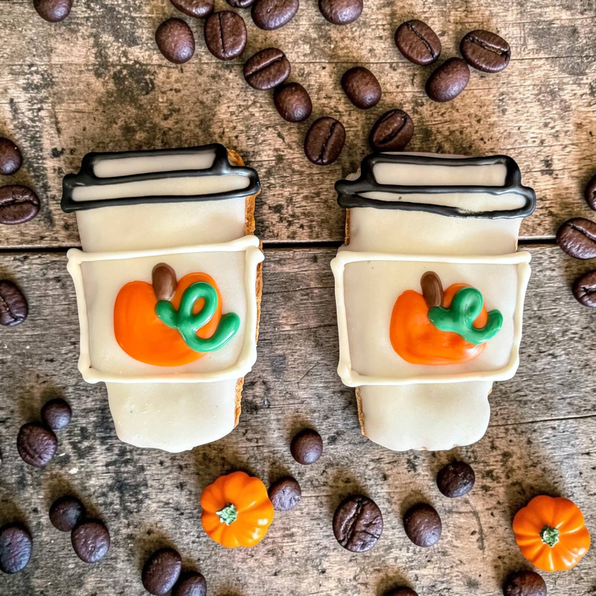 Two mini coffee-cup-shaped dog cookies with white decoration, black lid outlines, and orange pumpkins with green vines on a wooden surface.