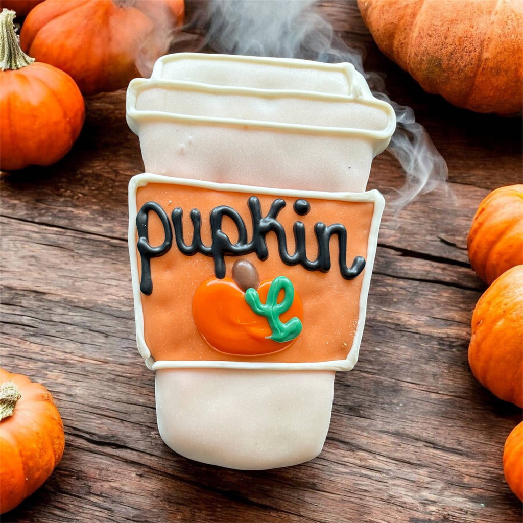 Coffee-cup-shaped dog cookie with white and orange decoration, black Pupkin lettering, and a small pumpkin design on a wooden surface.