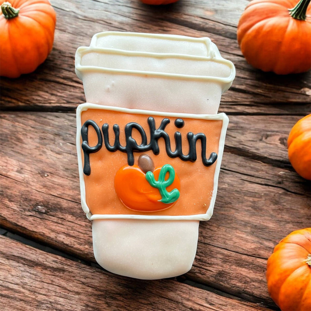 Coffee-cup-shaped dog cookie with white and orange decoration, black Pupkin lettering, and a small pumpkin design on a wooden surface.