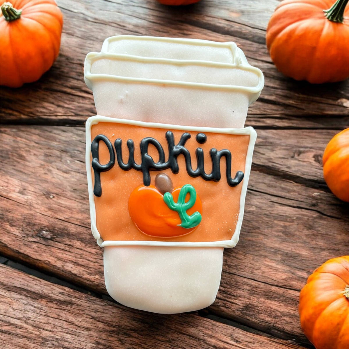 Coffee-cup-shaped dog cookie with white and orange decoration, black Pupkin lettering, and a small pumpkin design on a wooden surface.