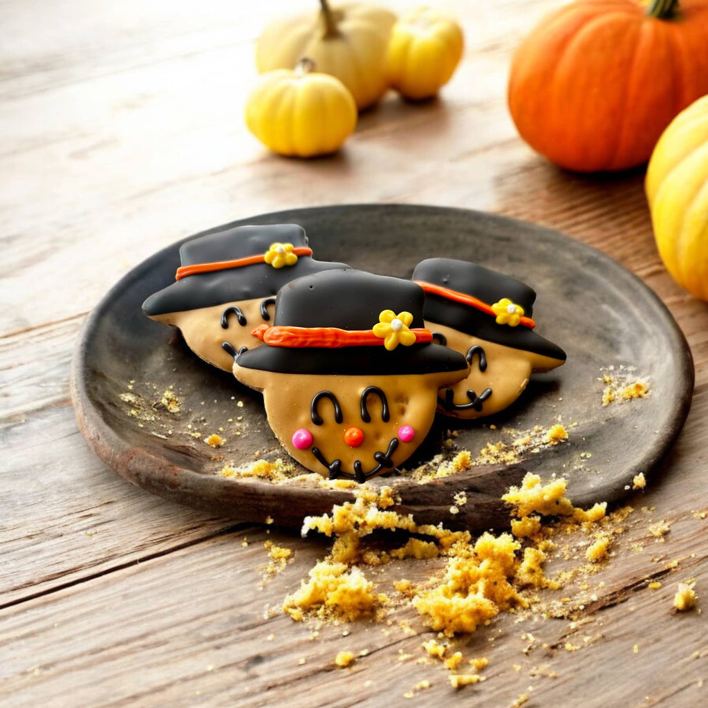 Three smiling scarecrow dog cookies with black hats, orange hatbands, and yellow flowers on a rustic plate.