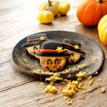 Three smiling scarecrow dog cookies with black hats, orange hatbands, and yellow flowers on a rustic plate.